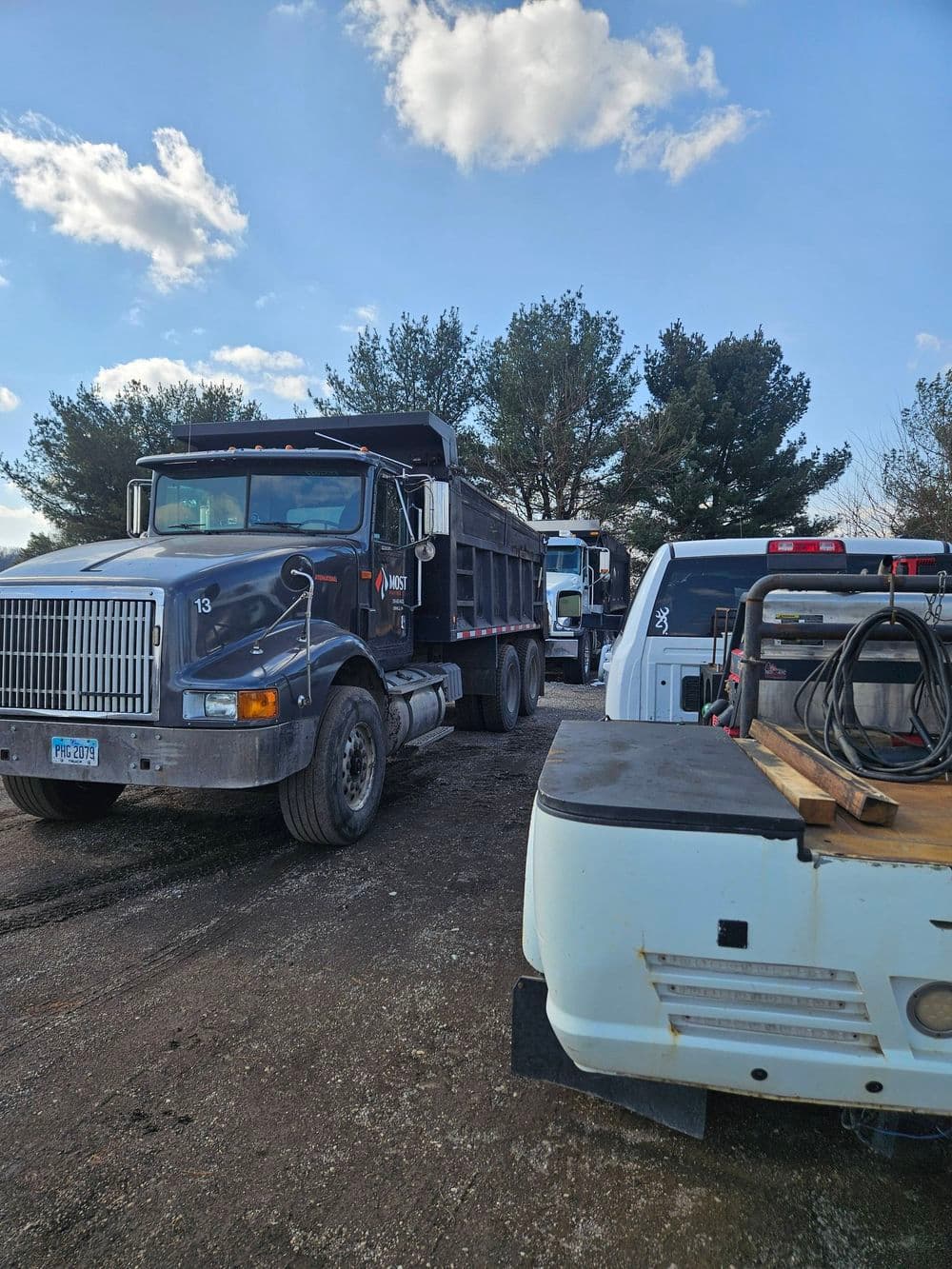 Dump truck and work vehicles lined up on a dirt road under a blue sky with clouds.